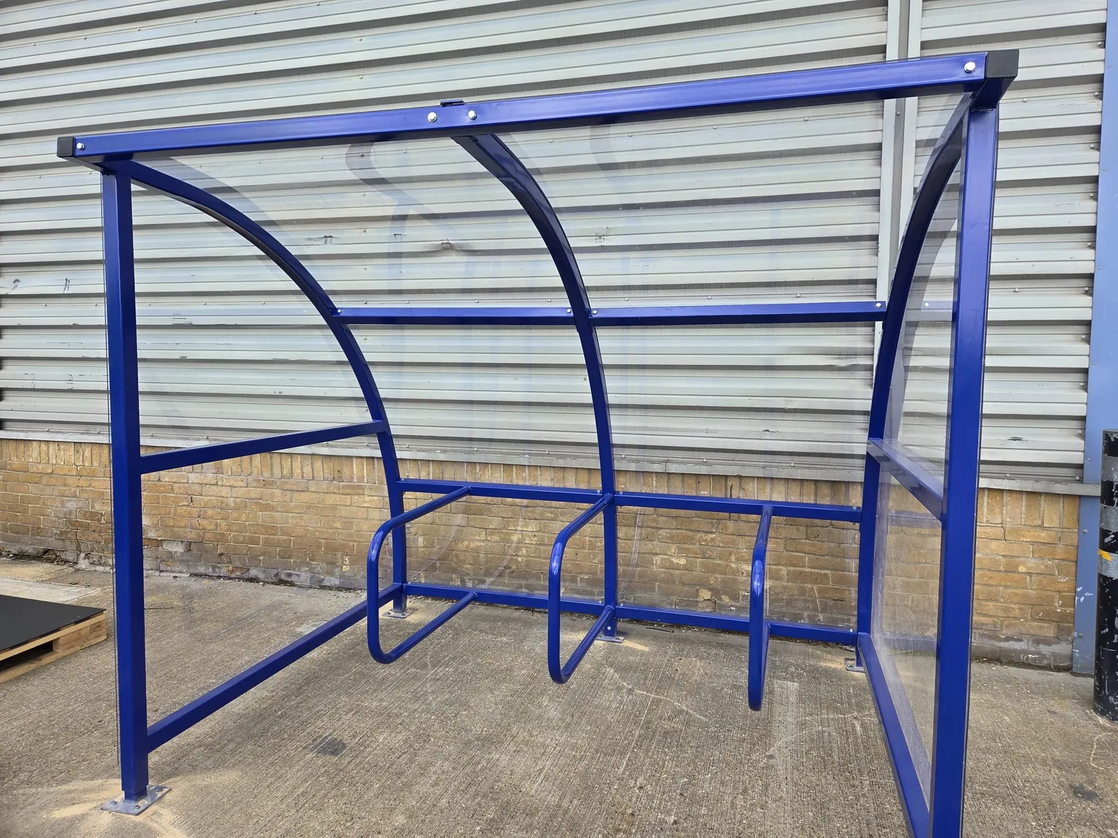 Blue steel-framed cycle shelter with curved polycarbonate roof and cycle hoops outside an industrial unit